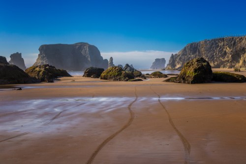 A serene beach scene with rocky formations, gentle waves, and clear blue skies, featuring tire tracks in the sand.