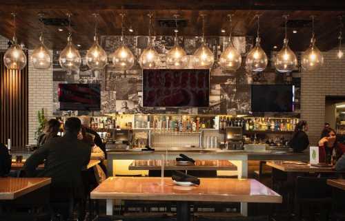 A modern restaurant interior featuring a bar with pendant lights, a large mural, and patrons enjoying their meals.