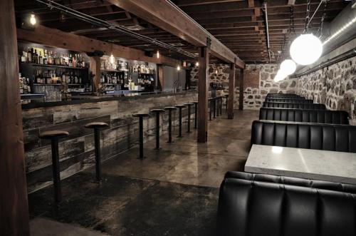 A dimly lit bar interior with wooden beams, a long bar counter, and black leather seating against stone walls.