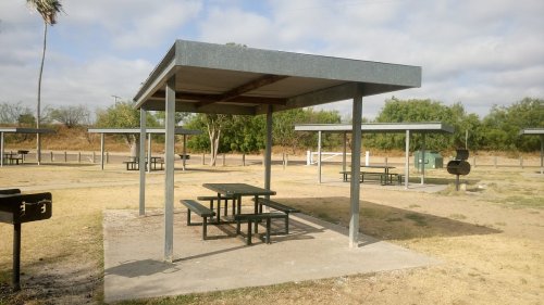 A picnic area with metal tables and a shelter, surrounded by grassy land and trees under a cloudy sky.