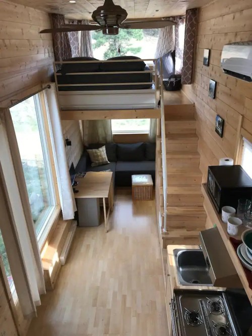Cozy tiny home interior with a lofted bed, living area, kitchen, and large windows for natural light.