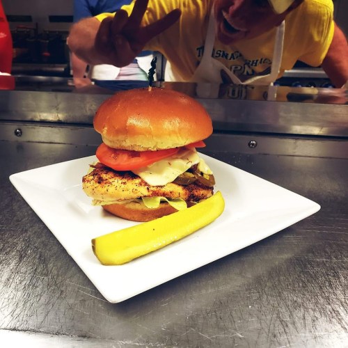 A delicious burger with grilled chicken, lettuce, tomato, and cheese on a plate, with a pickle and a person in the background.