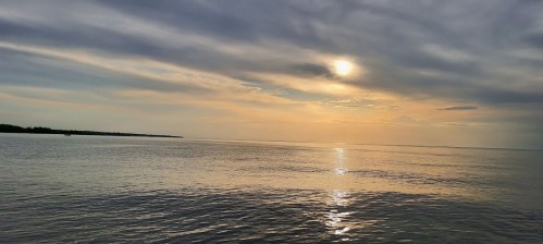 A serene seascape at sunset, with soft clouds reflecting on calm waters and a glowing sun on the horizon.