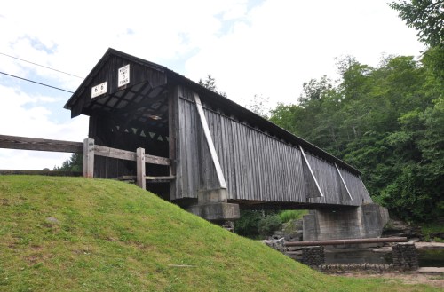 A wooden covered bridge stands over a grassy area, surrounded by trees under a partly cloudy sky.