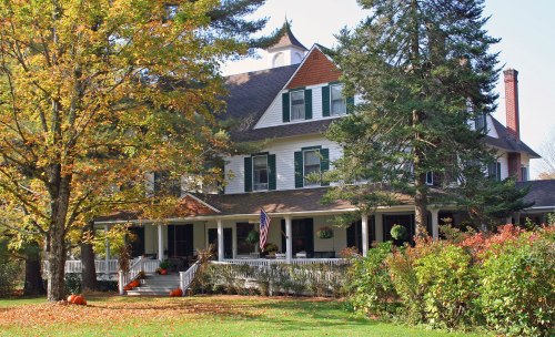 A large, charming house with a wraparound porch, surrounded by autumn trees and pumpkins in a vibrant fall setting.