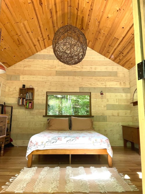 Cozy cabin interior with a wooden ceiling, a double bed, and a large window overlooking greenery.