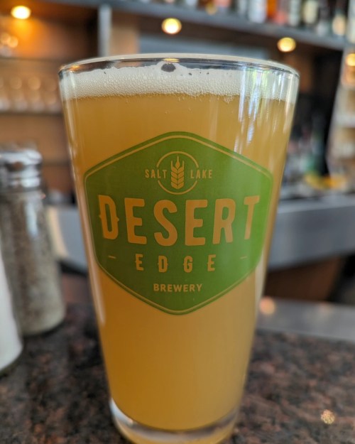 A close-up of a glass of beer with a green label reading "Desert Edge Brewery" on a bar countertop.