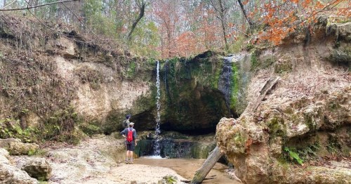 Hikers standing near a small waterfall in a rocky, forested area with a calm pool of water, on one the best waterfall hiking trails in Mississippi.