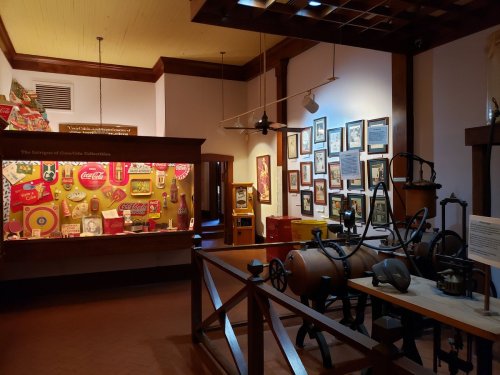A museum exhibit featuring vintage Coca-Cola memorabilia and historical artifacts displayed in a well-lit room.
