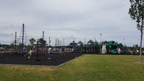 Children play on a playground with climbing structures and swings, surrounded by green grass and a cloudy sky.