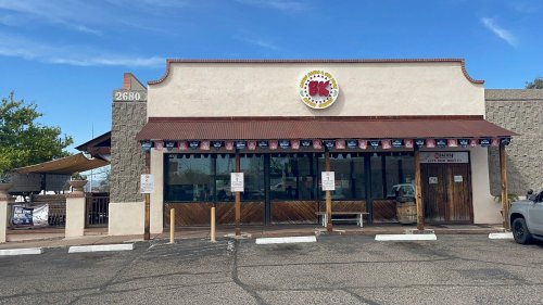 Exterior of a restaurant with a sign, awning, and flags, featuring a parking lot and clear blue sky.