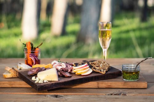 A wooden platter with assorted cheeses, meats, fruits, and bread, alongside a glass of sparkling wine and condiments.