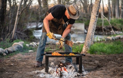 A person in a hat and apron grills food over an open fire in a natural outdoor setting.