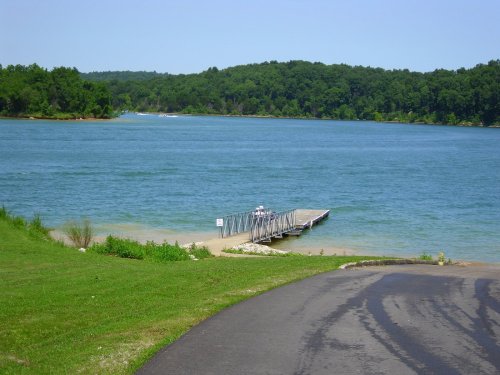 A serene lake view with a wooden dock, surrounded by green trees and a clear blue sky.