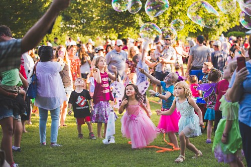 A lively outdoor scene with children in colorful outfits chasing bubbles among a crowd of people.