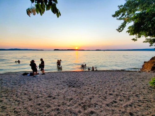 Sunset over a calm lake with people swimming and enjoying the beach on a warm evening.