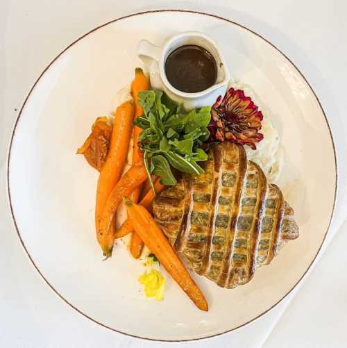 A plated dish featuring a pastry, carrots, mashed potatoes, and a small jug of sauce, garnished with greens and a flower.