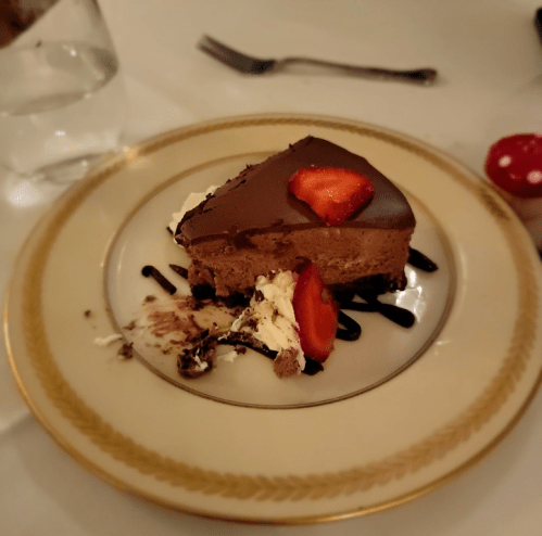 A slice of chocolate mousse cake topped with strawberries, served on a decorative plate with whipped cream and chocolate drizzle.