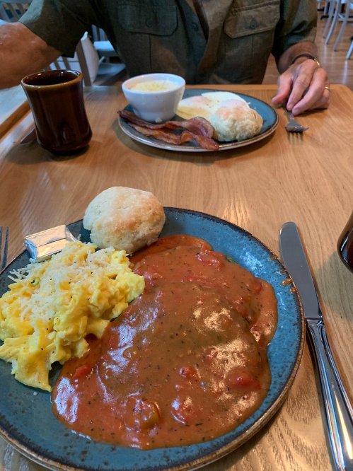 A plate of eggs, biscuits, and gravy with bacon, served alongside a bowl of soup on a wooden table.