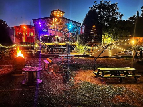A vibrant outdoor venue at night, featuring colorful lights, a fire pit, picnic tables, and a lively atmosphere.