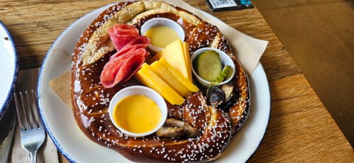 A large pretzel served with mustard, cheese, pickles, and slices of meat on a white plate.