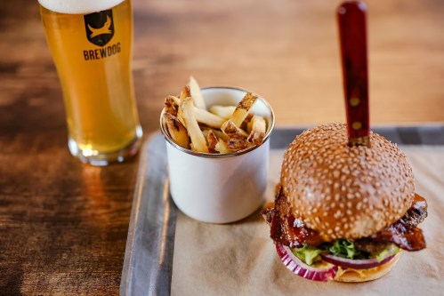 A sesame seed burger with bacon, lettuce, and onion, served with a side of fries and a glass of beer.