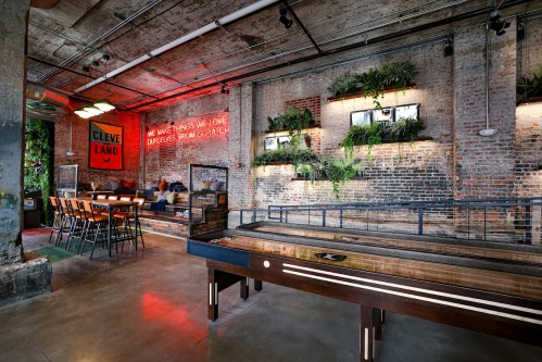 A stylish indoor space with a shuffleboard table, brick walls, greenery, and a dining area featuring modern seating.
