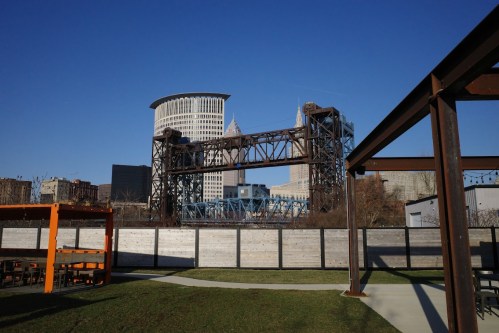 A view of a steel bridge with city skyscrapers in the background under a clear blue sky. Green grass in the foreground.