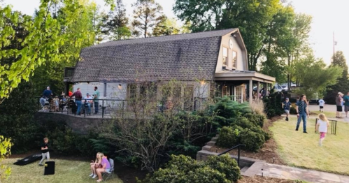 A cozy, two-story building with people gathered outside, surrounded by greenery and a lawn.