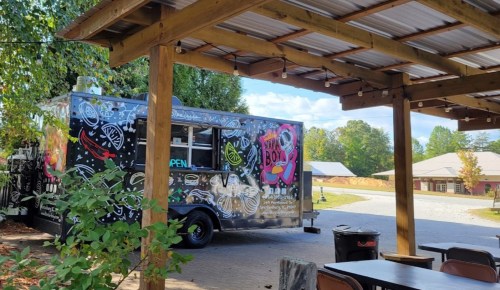 A colorful food truck parked under a wooden shelter, surrounded by trees and outdoor seating.