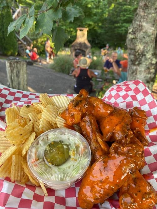 A plate of spicy chicken wings with chips and pickles, set outdoors with people enjoying the background.