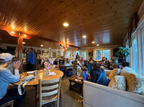A cozy café interior with wooden beams, people chatting, and a table with flowers, creating a warm, inviting atmosphere.