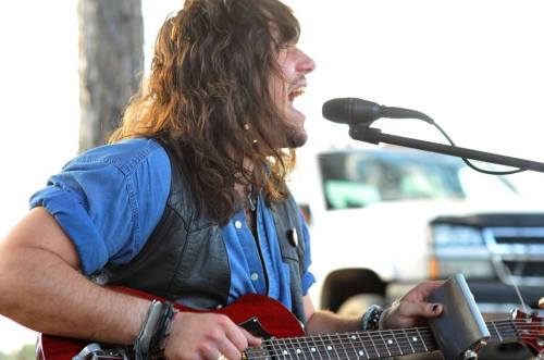 A musician with long hair passionately sings into a microphone while playing an acoustic guitar outdoors.