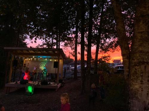 A band performs on an outdoor stage at sunset, surrounded by trees and an audience enjoying the music.