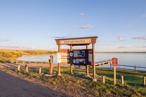 Signage at Brush Lake State Park with a calm lake and blue sky in the background, surrounded by grassy terrain.