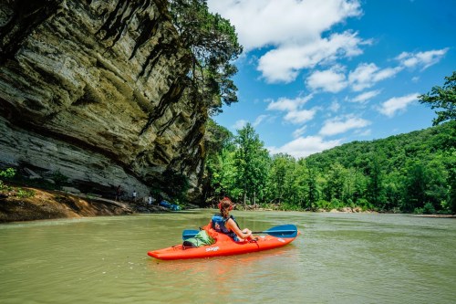 A person in a red kayak paddles on a calm river, surrounded by lush greenery and towering cliffs under a blue sky.