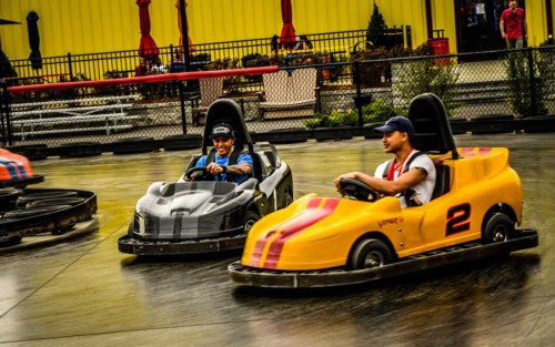 Two people race in go-karts on a track, one in a black car and the other in a yellow car with red stripes.