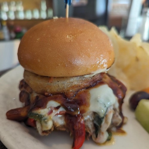 A close-up of a delicious slider topped with melted cheese, bacon, and a small blue toothpick, served with chips and fruit.
