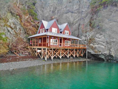 A charming red house on stilts by a rocky shore, surrounded by trees and clear turquoise water.