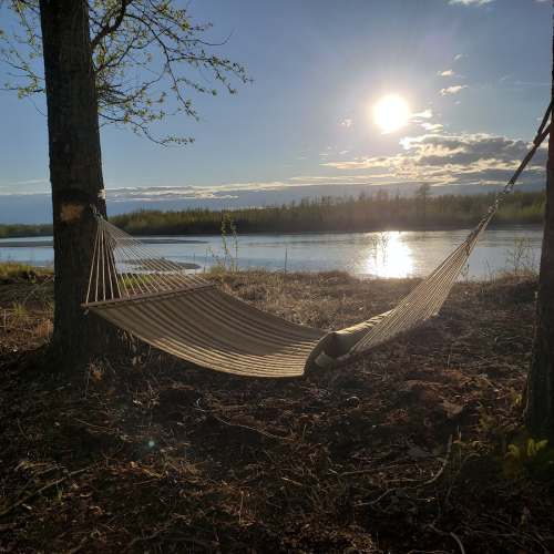 A hammock hangs between two trees by a river, with a sunset reflecting on the water and a serene landscape.