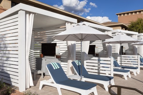 A poolside area with white cabanas, blue lounge chairs, and umbrellas under a clear blue sky.
