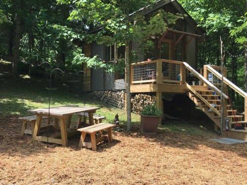 A cozy cabin surrounded by trees, featuring a wooden deck, picnic table, and a landscaped yard.