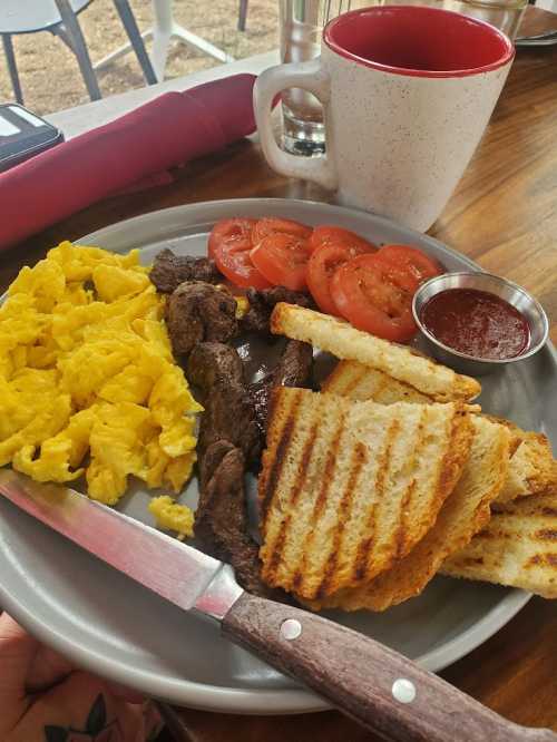 A plate with scrambled eggs, grilled steak, sliced tomatoes, toasted bread, and a small cup of sauce, with a coffee mug nearby.