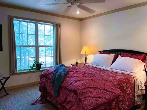Cozy bedroom with a large window, a neatly made bed with red bedding, and a lamp on a nightstand.