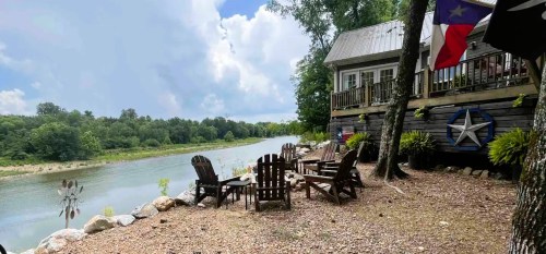 A serene riverside scene with wooden chairs, a house, and lush greenery under a partly cloudy sky.