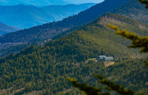 A solitary house nestled among lush green mountains, with rolling hills and distant blue peaks in the background.