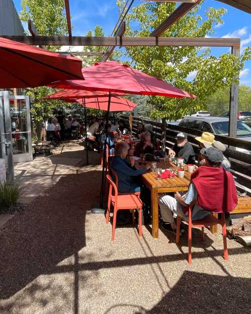 Outdoor dining area with red umbrellas, people seated at tables, enjoying a sunny day. Green trees in the background.