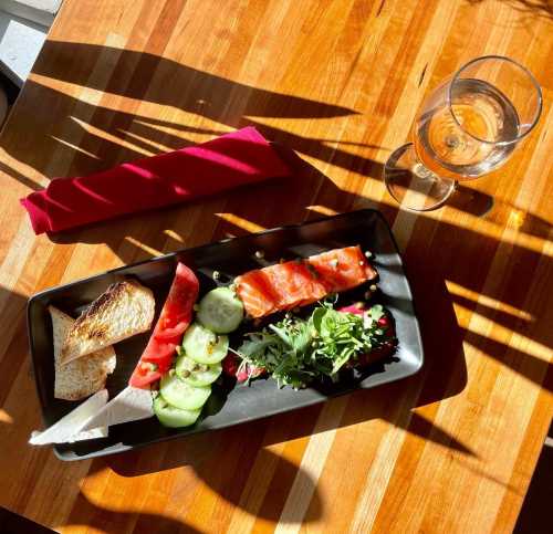 A black plate with salmon, cucumber, tomato, greens, and bread, alongside a glass of water on a wooden table.