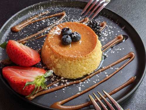 A creamy flan topped with caramel and berries, served on a black plate with chocolate drizzle and powdered sugar.