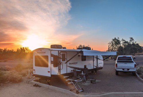 A camper trailer parked at sunset, with a truck nearby and chairs set up outside. Desert landscape in the background.